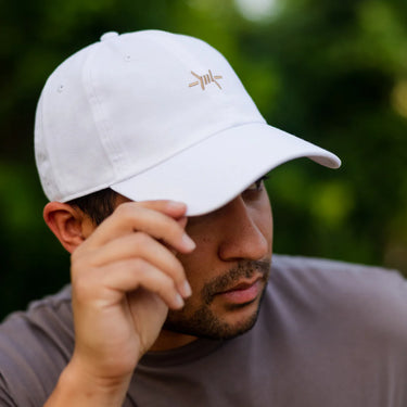 White Ball Cap at 6Whiskey six whisky Texas Standard. Men’s hats department-a close-up of a white cotton ball cap with a tan embroidered barbed wire logo on the front, curved brim, and structured crown, worn with a grey t-shirt against a green outdoor backdrop.
