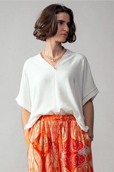 White Breezy V-Neck Top at 6Whiskey six whisky. A woman wears a relaxed white V-neck short-sleeve top with rolled cuffs, paired with a vibrant orange and red paisley print skirt.