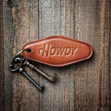 Howdy Keychain at 6Whiskey. Brown leather motel-style keychain embossed with “Howdy” in script, attached to a brass keyring with two vintage-style keys, resting on rustic wooden planks.