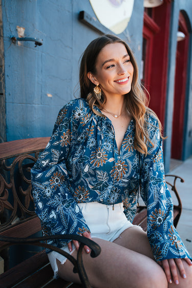 magnolia blouse at 6whiskey six whisky cleobella. A woman sits on a decorative metal bench in a small Texas town, wearing a blue floral long-sleeve blouse with gathered details and white high-rise shorts.
