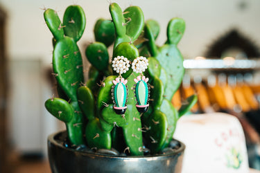 Cactus Drop Earrings at 6Whiskey. Pair of cactus-shaped drop earrings with green enamel, pink accents, and black pots, topped with faux pearl cluster studs, displayed on a vibrant potted cactus