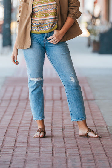 Hailey High-Rise Jeans at 6Whiskey six whisky. Women’s jeans department-a model wears light wash, high-rise straight-leg jeans with subtle knee distressing and a raw hem, styled with brown sandals, a patterned knit tank, and a tan utility jacket on a brick sidewalk.