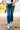 Audrey Cuff Straight Jeans Denim at 6Whiskey six whisky. Women’s jeans department-close-up side view of a model wearing medium wash, high-rise straight-leg jeans with stitched front seams and wide rolled cuffs, styled with a white tee and pale blue ankle boots against a yellow mural wall.