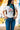 Audrey Cuff Straight Jeans Denim at 6Whiskey six whisky. Women’s jeans department-close-up side view of a model wearing medium wash, high-rise straight-leg jeans with stitched front seams and wide rolled cuffs, styled with a white tee and pale blue ankle boots against a yellow mural wall.