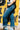 Audrey Cuff Straight Jeans Denim at 6Whiskey six whisky. Women’s jeans department-close-up side view of a model wearing medium wash, high-rise straight-leg jeans with stitched front seams and wide rolled cuffs, styled with a white tee and pale blue ankle boots against a yellow mural wall