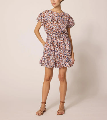 Aurora Mini Dress at 6Whiskey six whisky Cleobella. A woman wearing a short, flutter-sleeve mini dress with a high neckline, removable waist sash, and an allover floral print in earthy tones; the dress features a ruffled hem and side pockets, styled with brown ankle-strap sandals.