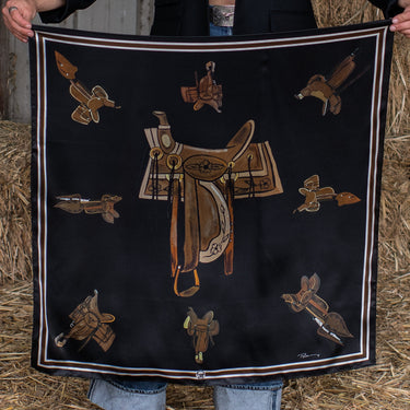Take a seat silk scarf by Fringe Scarves at 6Whiskey, six whisky, equestrian theme, saddle, horse saddles, white, black, brown, burgundy, maroon, square, soft, comfortable, breathable, year-round, must-have, eye-catching, classic western, timeless, elevated, front view. Scarf with equestrian-themed design held by a person in a rustic setting. Texas A&M gameday, game day, A&M, Aggie style, western aggie style for gamedays, football gameday style SEC, wear to Kyle field or tailgates.