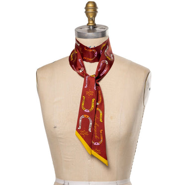 "Horseshoe Twilly" – a hand-illustrated, 100% silk twilly scarf in a rich maroon hue, featuring golden horseshoes, equestrian ribbon details, and the mantra "Giddy Up." Styled on a dress form, the scarf showcases its versatile design, perfect for wearing as a necktie, hair accessory, or bag accent. Finished with golden-yellow edges for a bold contrast.