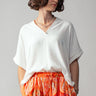 White Breezy V-Neck Top at 6Whiskey six whisky. A woman wears a relaxed white V-neck short-sleeve top with rolled cuffs, paired with a vibrant orange and red paisley print skirt.