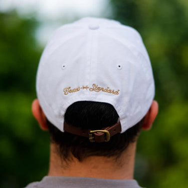 White Ball Cap at 6Whiskey six whisky Texas Standard. Men’s hats department-a close-up of a white cotton ball cap with a tan embroidered barbed wire logo on the front, curved brim, and structured crown, worn with a grey t-shirt against a green outdoor backdrop.