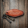 Howdy Keychain at 6Whiskey. Brown leather motel-style keychain embossed with “Howdy” in script, attached to a brass keyring with two vintage-style keys, resting on rustic wooden planks.