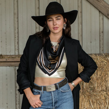 Take a seat silk scarf by Fringe Scarves at 6Whiskey, six whisky, equestrian theme, saddle, horse saddles, white, black, brown, burgundy, maroon, square, soft, comfortable, breathable, year-round, must-have, eye-catching, classic western, timeless, elevated, front view. Scarf with equestrian-themed design worn by a woman in a rustic setting. Texas A&M gameday, game day, A&M, Aggie style, western aggie style for gamedays, football gameday style SEC, wear to Kyle field or tailgates.