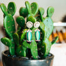 Cactus Drop Earrings at 6Whiskey. Pair of cactus-shaped drop earrings with green enamel, pink accents, and black pots, topped with faux pearl cluster studs, displayed on a vibrant potted cactus