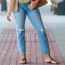 Hailey High-Rise Jeans at 6Whiskey six whisky. Women’s jeans department-a model wears light wash, high-rise straight-leg jeans with subtle knee distressing and a raw hem, styled with brown sandals, a patterned knit tank, and a tan utility jacket on a brick sidewalk.