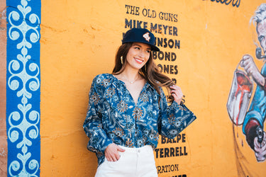 magnolia blouse at 6whiskey six whisky cleobella. A woman sits on a decorative metal bench in a small Texas town, wearing a blue floral long-sleeve blouse with gathered details and white high-rise shorts.