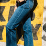 Audrey Cuff Straight Jeans Denim at 6Whiskey six whisky. Women’s jeans department-close-up side view of a model wearing medium wash, high-rise straight-leg jeans with stitched front seams and wide rolled cuffs, styled with a white tee and pale blue ankle boots against a yellow mural wall