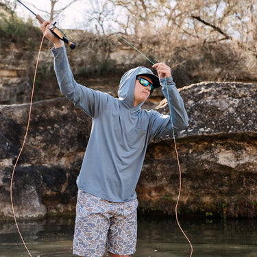 Man wearing Texas Standard Brazos Hoodie in Bayou Blue while fly fishing, showcasing UPF 50+ sun protection, moisture-wicking performance, and breathable stretch fabric – available at 6Whiskey, Texas lifestyle clothing store