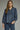 Denim darling blouse at 6whiskey, six whisky, woman wearing a blue denim blouse with jeans, ideal for fall game day or tailgate looks at texas a&m, aggies, SEC, gameday outift inspo, ruffle detail blouse with buttons and self tie, elasticated gathered long sleeve blouse, front view, football game outfit, watch party look, game day to weekday look. Pair with maroon or burgundy accessories and sneakers, sandals, or boots. Texas A&M casual chic aggie style.