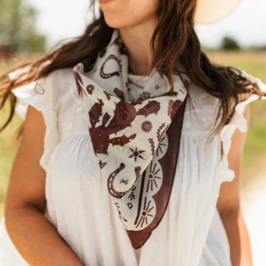 Down south bandana at 6whiskey, six whisky, Texas A&M maroon burgundy western icon graphic, maroon and white, soft, lightweight, breathable, comfortable, accessory, scarf, bandana, gameday, tailgating, football, SEC, western, casual. Front view. Woman wearing white blouse and bandana. Tie on neck, wrist, beltloops, or on purse. Aggie style and gameday outfit inspo. Horseshoe, boots, bucking horse and cowboy and cactus.