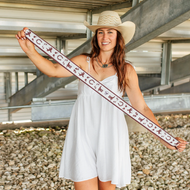 Down south twilly at 6whiskey, six whisky, Texas A&M maroon burgundy western icon graphic, maroon and white, soft, lightweight, breathable, comfortable, accessory, scarf, twilly, ribbon, gameday, tailgating, football, SEC, western, casual. Front view. Woman wearing white dress holding twilly. Tie on neck, wrist, beltloops, or on purse. Aggie style and gameday outfit inspo. Horseshoe, boots, bucking horse and cowboy.