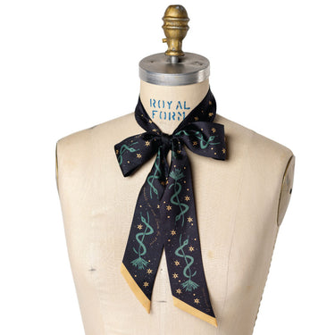 Lasso the Stars Twilly – a luxurious, hand-illustrated 100% silk twilly scarf with a midnight black background, adorned with golden stars and deep teal swirling lasso motifs. The design embodies a celestial Western theme, symbolizing adventure and limitless possibilities. With gold-accented edges and a sleek, elongated shape (1.5" x 55"), this versatile accessory can be styled as a necktie, headband, or bag accent, adding a touch of cosmic cowgirl charm to any outfit.