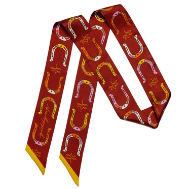 "Horseshoe Twilly" – a hand-illustrated, 100% silk twilly scarf in a rich maroon hue, featuring golden horseshoes, equestrian ribbon details, and the mantra "Giddy Up." Styled on a dress form, the scarf showcases its versatile design, perfect for wearing as a necktie, hair accessory, or bag accent. Finished with golden-yellow edges for a bold contrast.